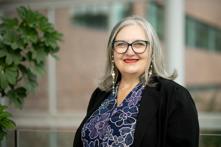 A woman with grey hair and glasses. She is wearing a blue floral shirt with a dark jacket. In the background is the blurry side of a brick building.